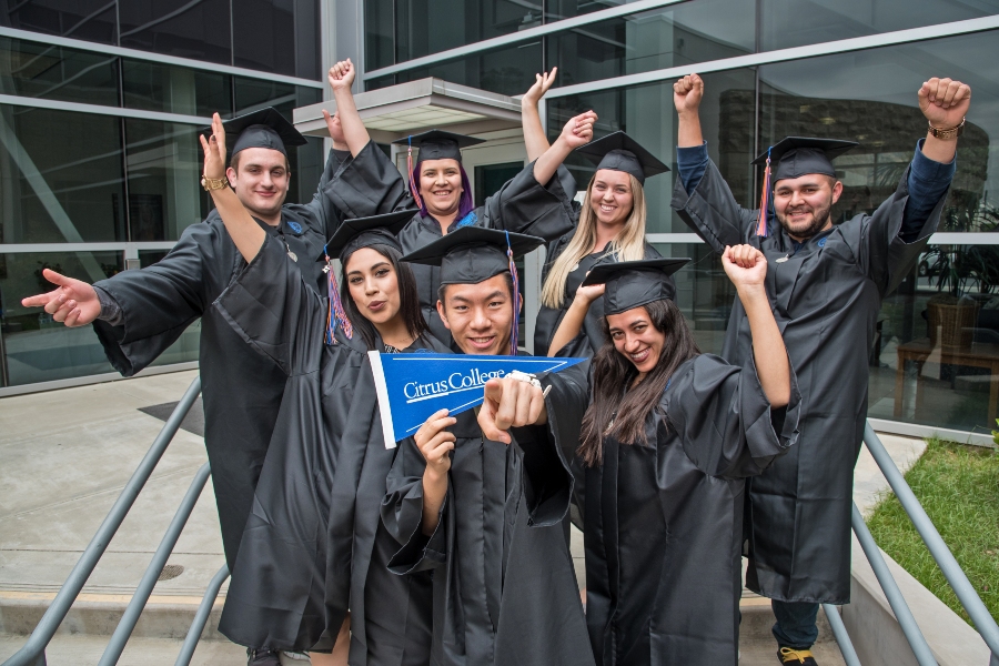 seven citrus college graduates in caps and gowns celebrate outside a campus building raising their arms and holding a citrus college pennant