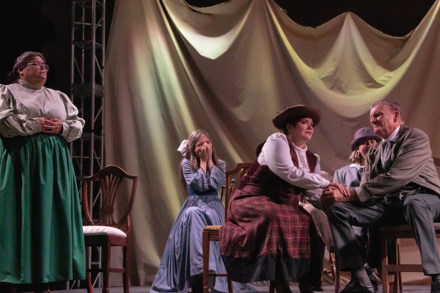 actors in period costumes perform a scene from our town during an outdoor citrus college theatre arts production, with several cast members gathered onstage beneath draped fabric scenery