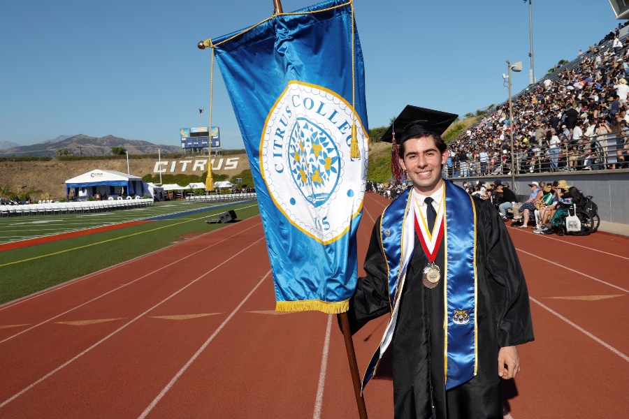 student trustee aj hernandez in cap and gown holds a citrus college banner on the stadium track during commencement, with medals around his neck and graduates, guests and stadium commencement set up in the background.