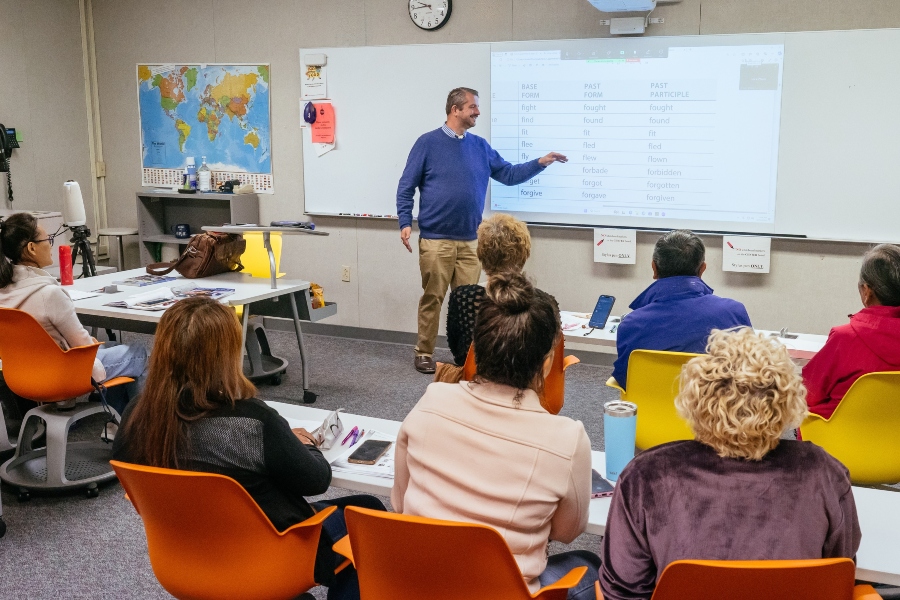 instructor stands at the front of a classroom pointing to a projected lesson on verb forms while adult students sit at tables facing the presentation