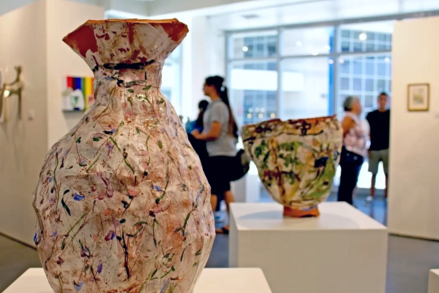 large textured ceramic vessel displayed on a pedestal in an art gallery, with another vessel and visitors in the background