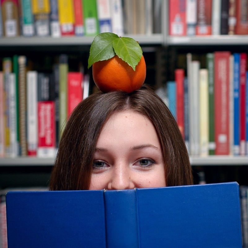 student holding an open book in a library with an orange balanced on their head, symbolizing learning and academic achievement
