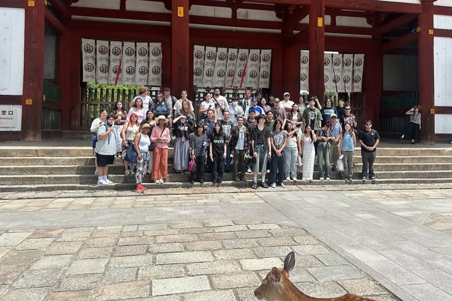 citrus college study abroad students and instructors pose on the steps of a traditional japanese temple or shrine, with red wooden columns and hanging banners behind them and a deer crossing the foreground