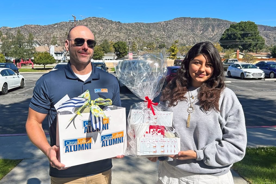 a man and woman stand outdoors with mountains in the background, holding citrus college alumni gift boxes and a wrapped chick-fil-a gift basket as part of a firefighter appreciation visit