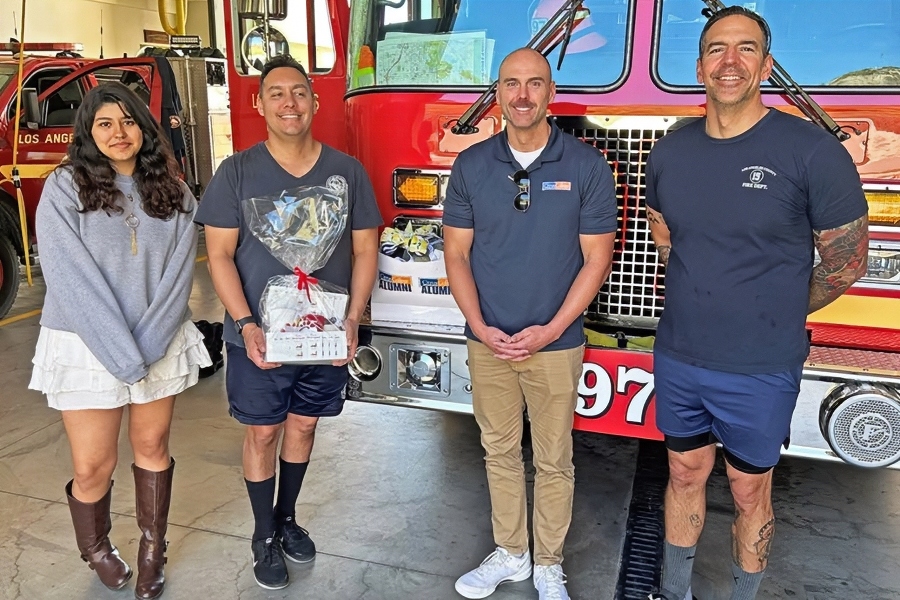a woman and three firefighters stand inside los angeles county fire department station 97 in front of a red fire engine; one firefighter holds a wrapped appreciation gift basket, with citrus college alumni gifts visible behind them