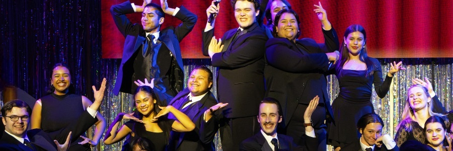 performers in formal black attire strike playful poses together onstage against a shimmering backdrop during the anniversary gala