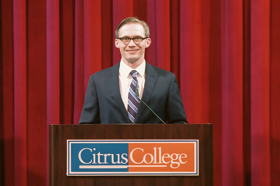 a man in a suit speaks from a citrus college podium in front of red stage curtains
