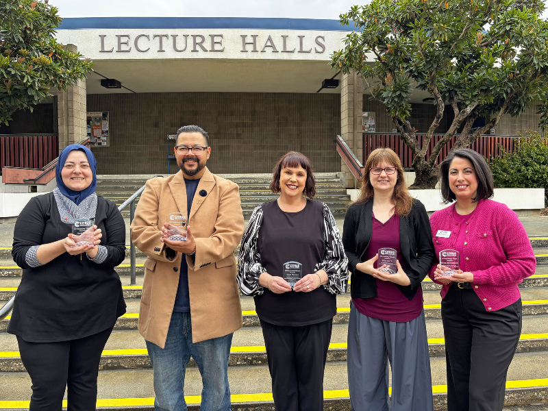 five staff members standing on the steps outside the lecture halls building, each holding a clear award plaque and smiling at the camera, with trees in the background