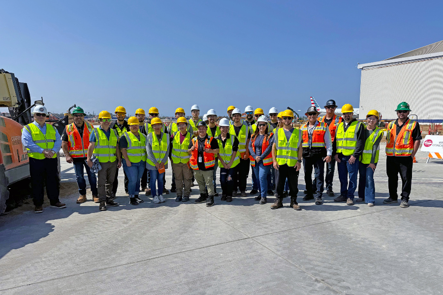 construction management students, faculty and industry professionals wearing hard hats and safety vests stand together at an active construction site during a program visit