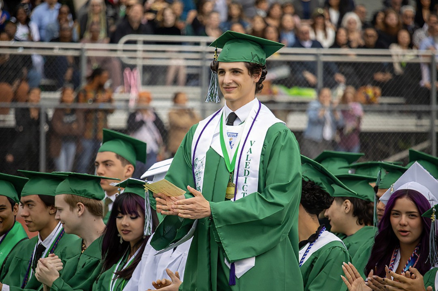 high school valedictorian in green cap and gown stands and applauds during graduation ceremony surrounded by classmates in stadium seating