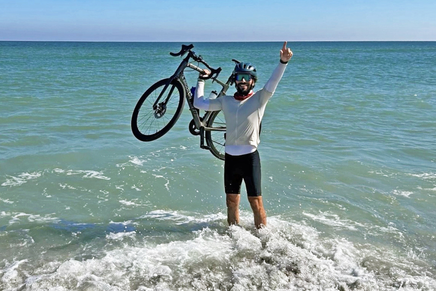 cyclist standing in ocean surf while holding a bicycle overhead after completing a cross-country ride