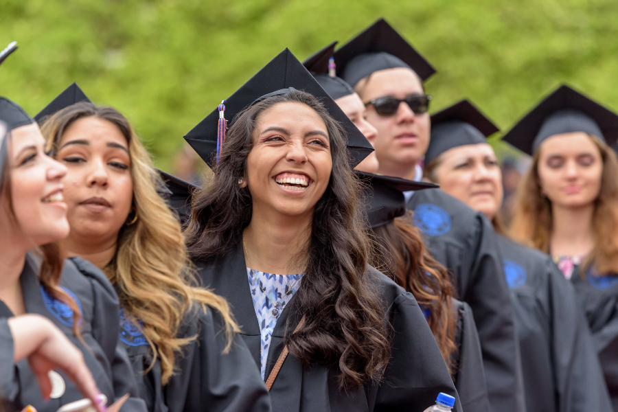 smiling graduates in black caps and gowns stand together during a commencement ceremony, with one graduate in the center laughing as classmates gather around her outdoors against a green backdrop
