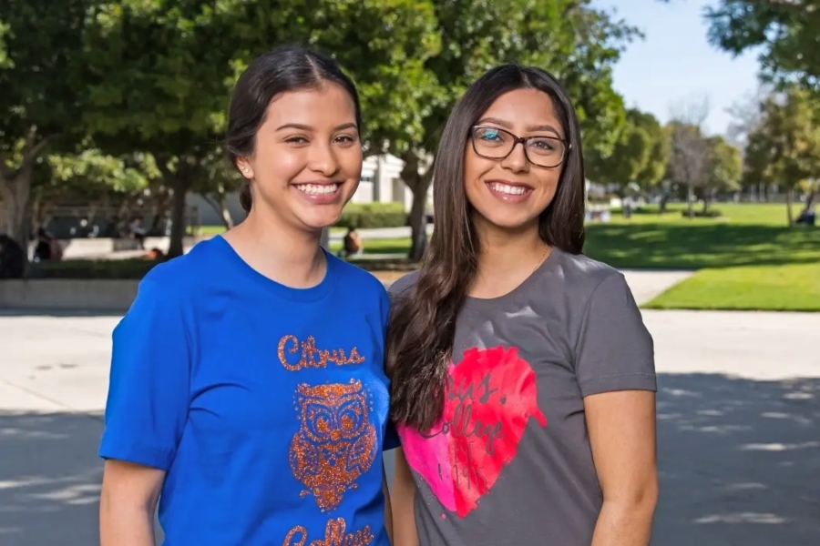 two smiling women wearing citrus college tee shirts stand together outdoors on campus, with trees and a grassy lawn in the background