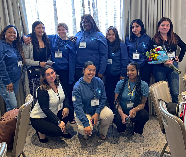 ten students and staff posing indoors at a regional conference, wearing matching blue hoodies and name badges, smiling together in a meeting room with chairs and tables, one person holding a bouquet of flowers