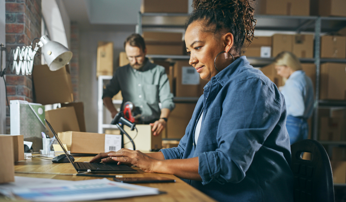 woman works on a laptop at a desk while two coworkers handle packages in a warehouse workspace