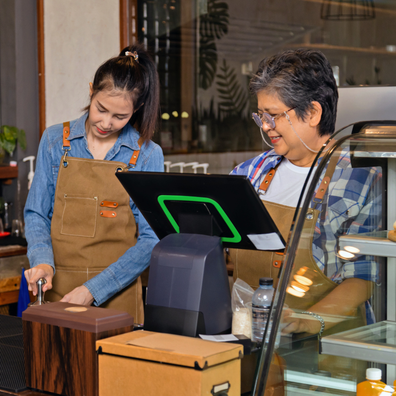 two women wearing aprons work together behind a café counter, one preparing a drink while the other uses a point-of-sale register beside a glass display case and countertop equipment