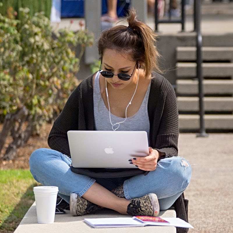 young woman sitting cross-legged outdoors on a low concrete ledge, working on a laptop