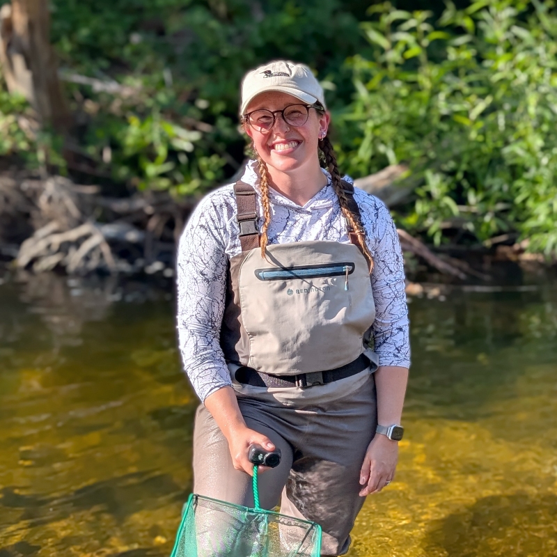 a smiling Emily Correia-Lambton wearing glasses, a cap and chest waders while standing outdoors in a stream