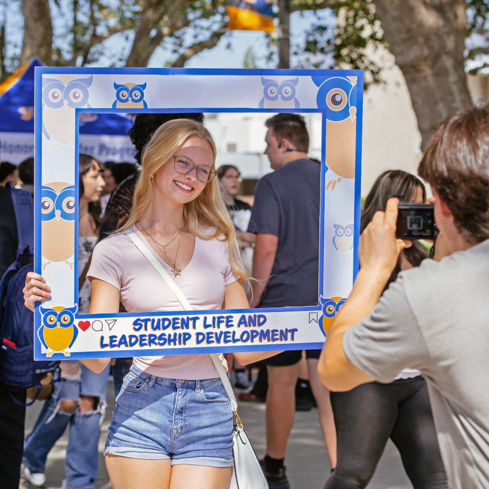 student poses inside an owl-themed student life and leadership development frame while another person takes her photo at an outdoor campus event