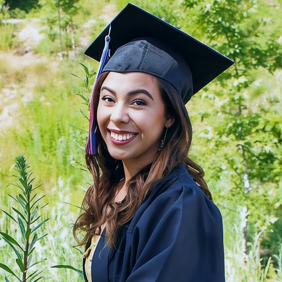 a smiling kaitlyn espinose wearing a black cap and gown poses outdoors on a deck surrounded by green plants and trees