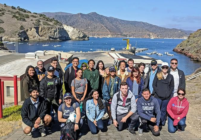 students and staff pose together overlooking a coastal harbor with boats, rocky cliffs and mountains in the background