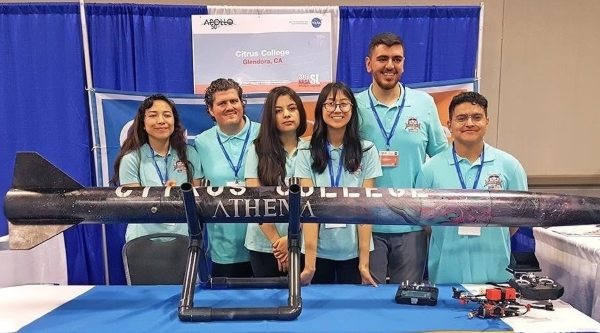 six rocket owls students pose behind a large model rocket at a conference exhibit booth