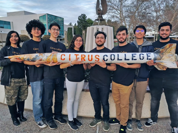 eight citrus college students stand together outdoors on campus, holding a large model rocket labeled citrus college