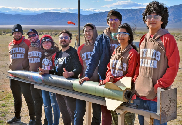 rocket owls students and faculty pose beside a student-built rocket at a desert launch site, with mountains visible in the distance