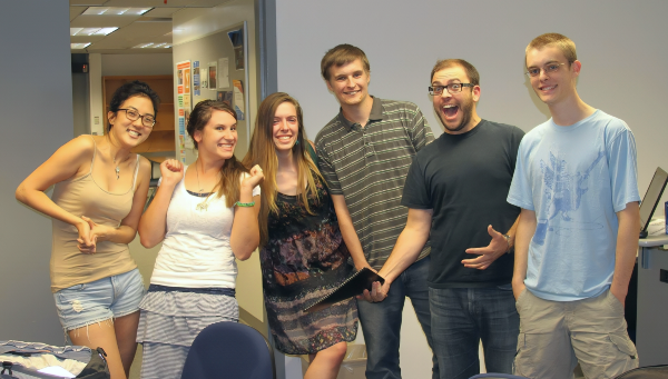 six students pose playfully together in a classroom or lab, smiling at the camera with desks, computers and bulletin boards visible in the background