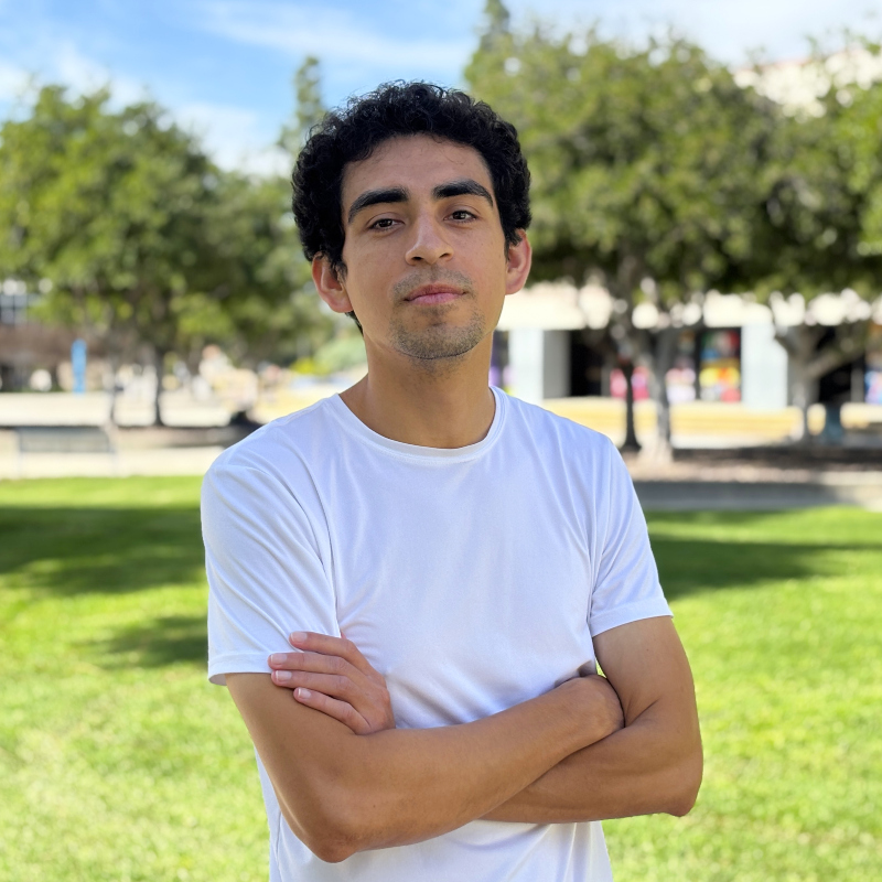 Majin Perez standing outdoors on a campus lawn with arms crossed wearing a white t shirt trees and buildings in the backgroundnstructors Amanda Keller Konya and Kim Toth.