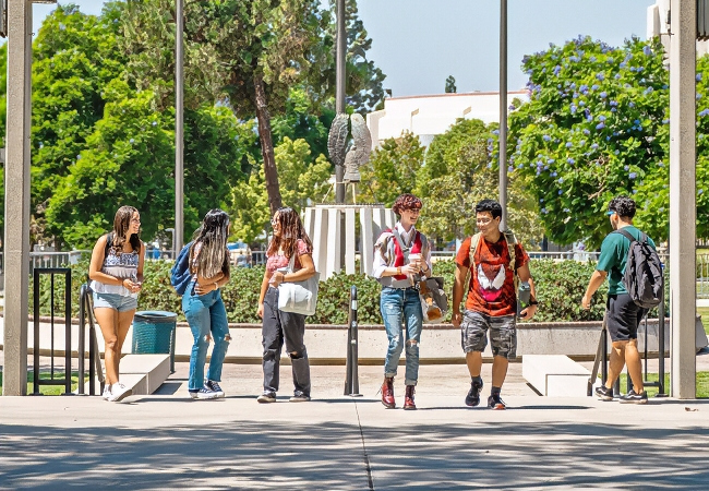 group of college students walking and talking on a sunny campus walkway near a central courtyard owl statue, carrying backpacks and enjoying a relaxed outdoor setting